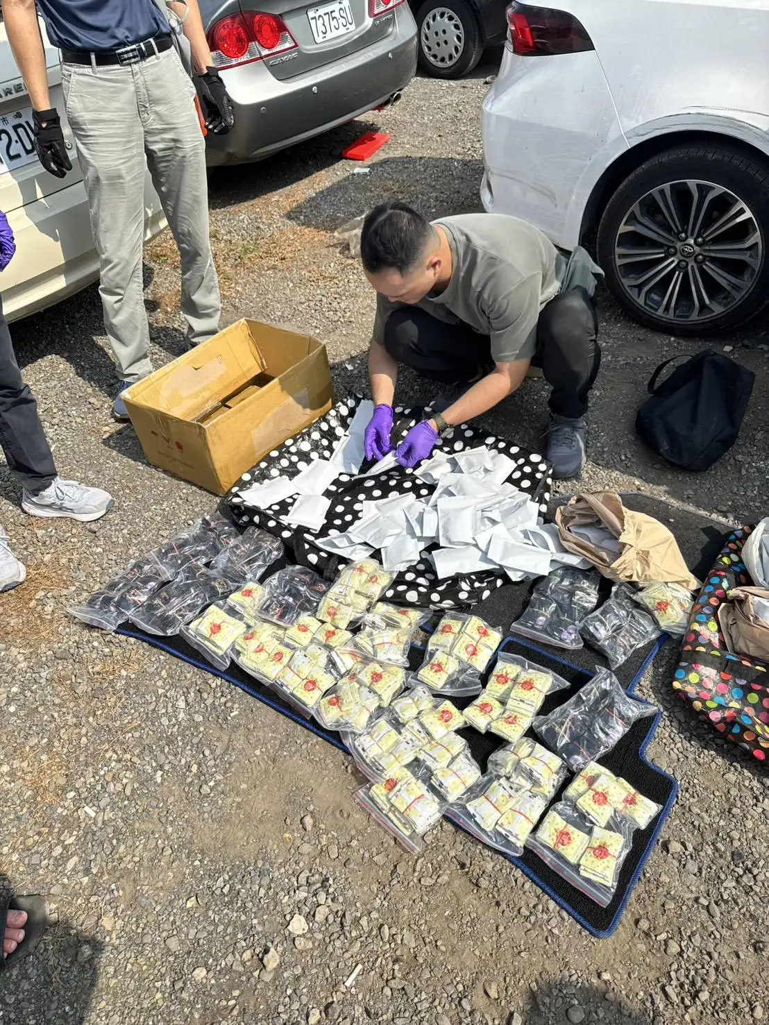 專案小組於空地現場破獲移動式毒庫，幹員逐一清點查扣之數千包毒品咖啡包、彩虹菸及相關包裝證物。(圖 / 張振興翻攝)